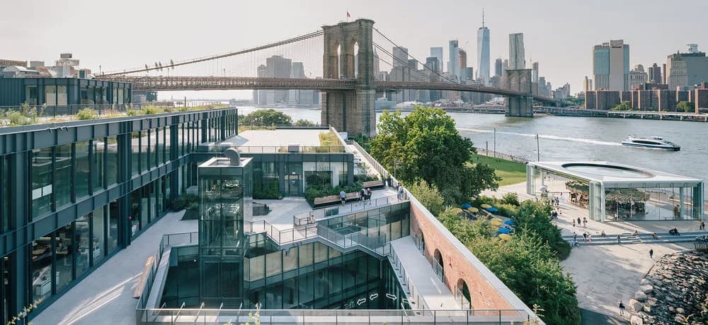 Modern glass-and-steel waterfront building beneath the Brooklyn Bridge with the Manhattan skyline beyond